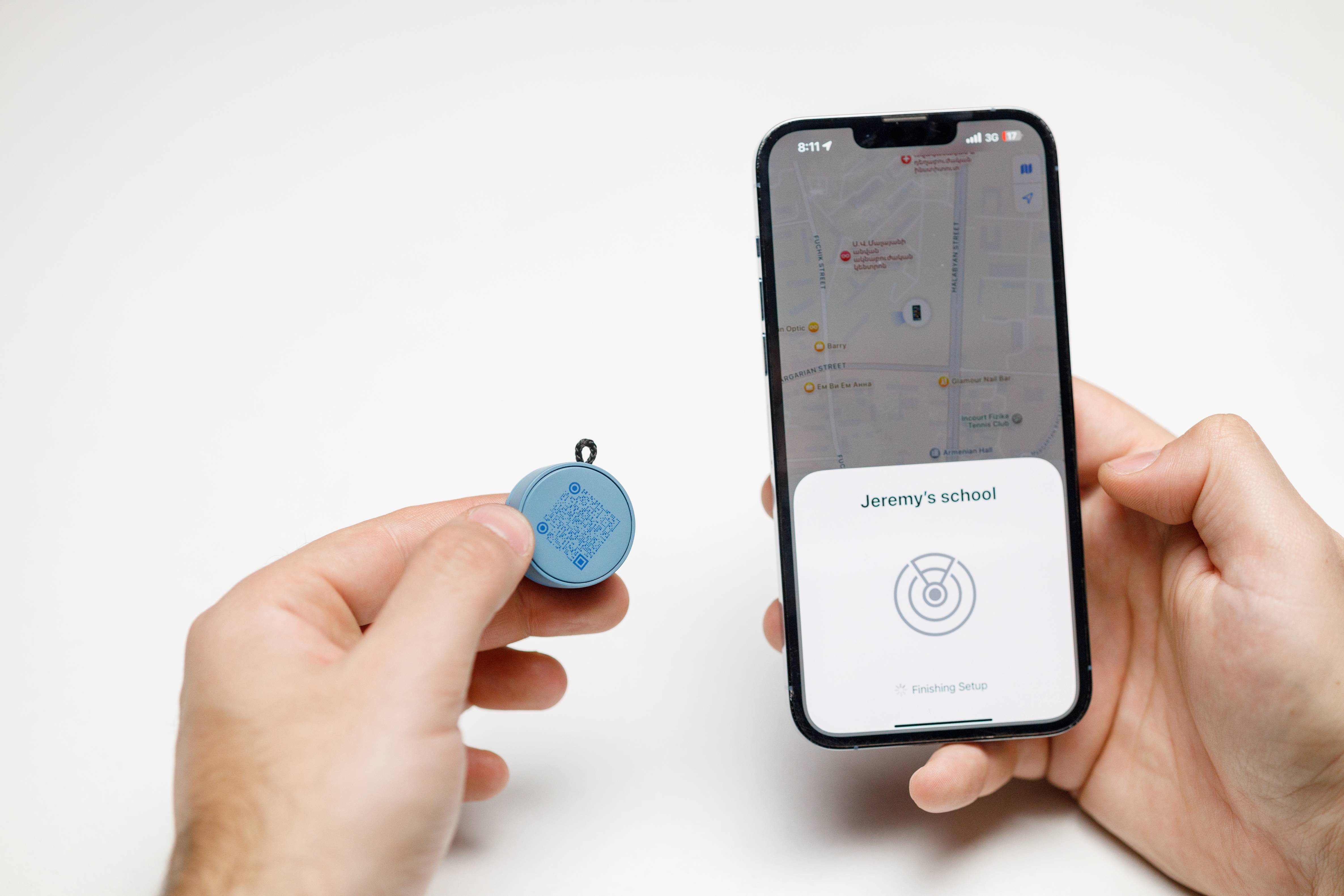 Two hands holding a blue Roam Smart Tracker and an iPhone showing Apple Find My app setup screen for adding the tracker