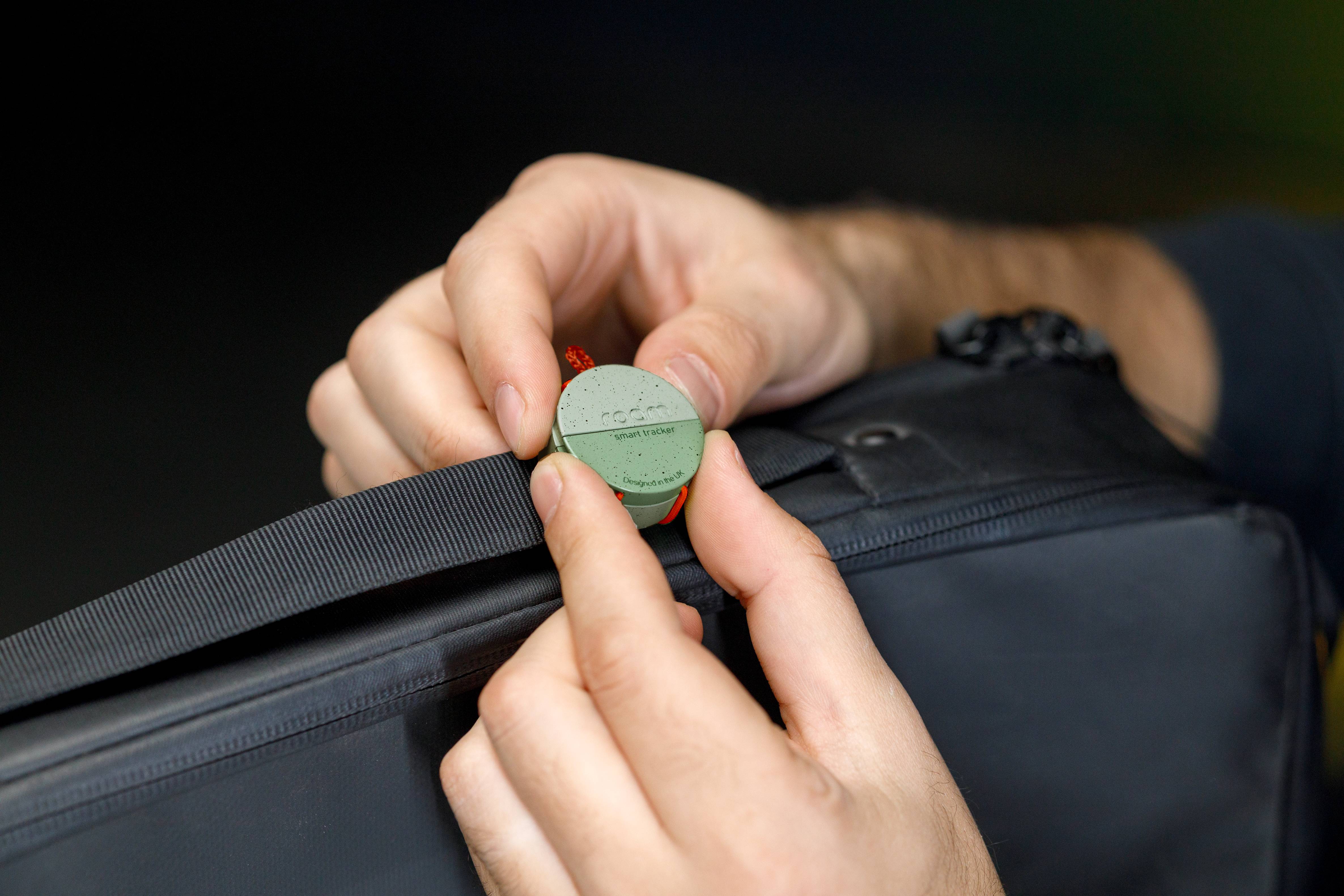 Hands attaching a green Roam Smart Tracker to the strap of a dark backpack illustrating how smart trackers compare to GPS trackers for everyday use