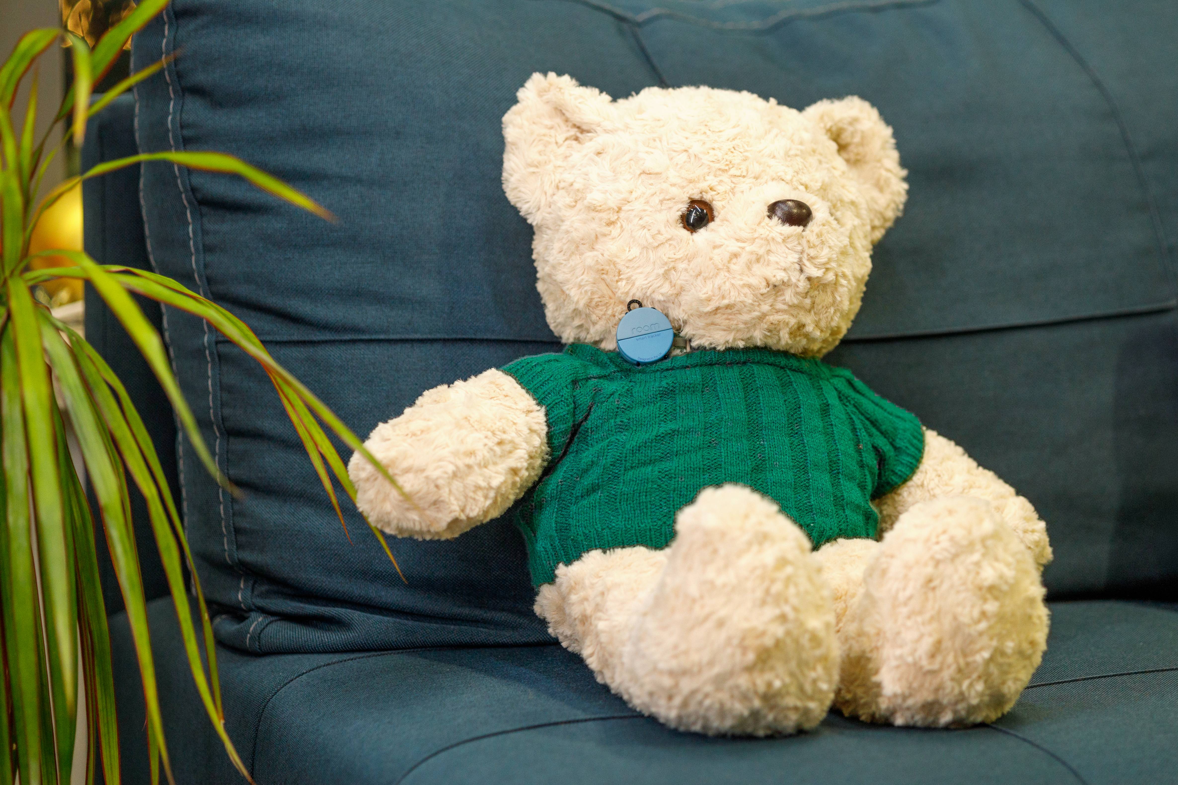 Teddy bear wearing green knit sweater sitting on blue couch with Roam Smart Tracker attached showing how to track kids belongings and soft toys