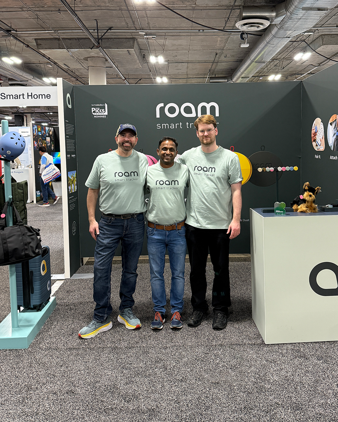 Roam smart tracker team at CES trade show booth wearing Roam branded t-shirts with smart tracker product display and All Colors lineup in background
